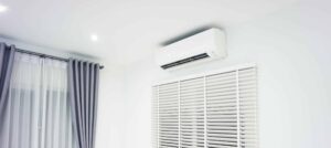 Modern white ductless mini-split air conditioning unit mounted on wall above shutters in a bright room.