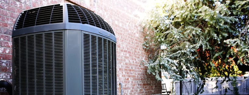 Gray cylindrical air conditioning unit installed against a brick wall with green ivy nearby.