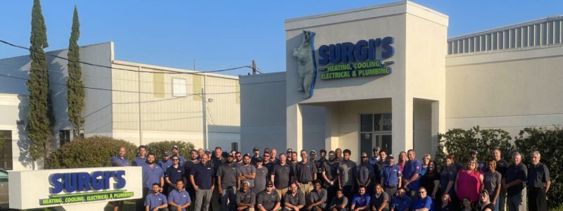 Large group of HVAC and plumbing technicians in uniforms standing together outside Surgi's Heating, Cooling, Electrical & Plumbing storefront in Louisiana.